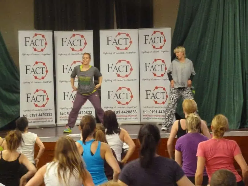 Two instructors leading a dance fitness class on stage in front of FACT charity banners.