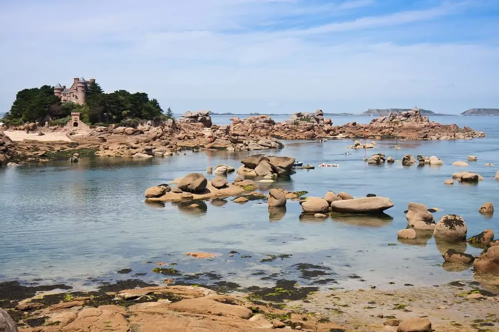 Rocky coastline in Brittany with pink granite rock formations, calm blue water, and a historic castle nestled on a small island.