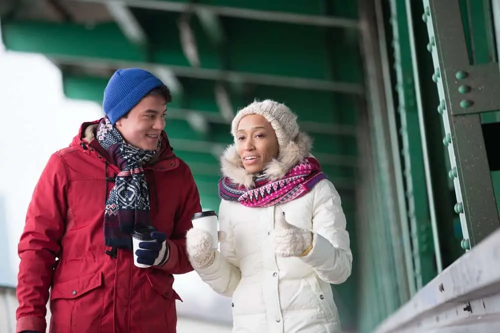 A couple in winter coats, beanies, and scarves holding coffee cups and walking together outdoors.