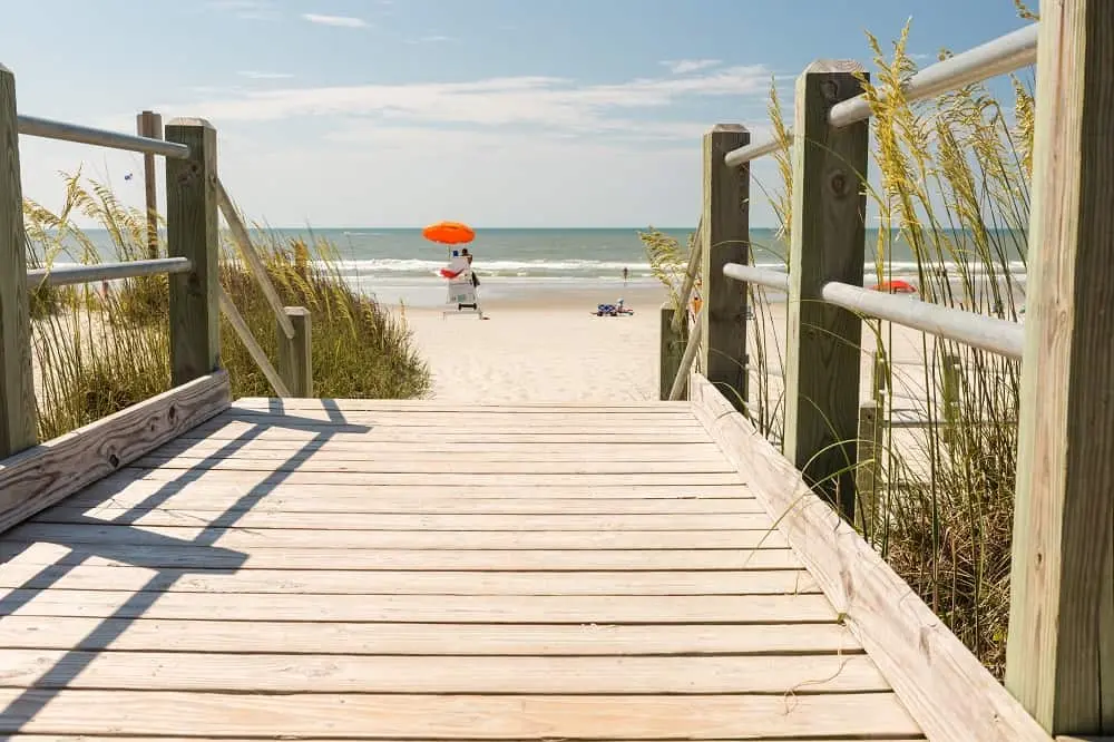 Wooden boardwalk leading to a sandy beach with an orange umbrella and ocean views.