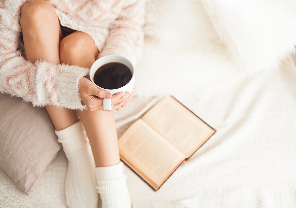 A woman sat on the bed drinking coffee and reading a book