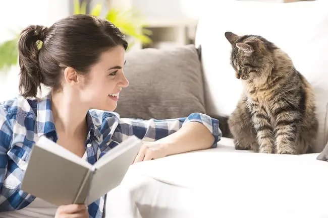 A smiling woman holding an open book and looking up at a fluffy tabby cat sitting on a white couch.