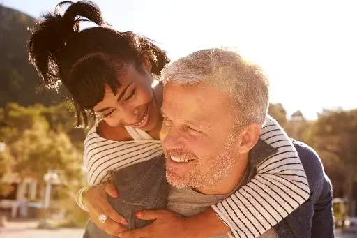 A smiling man giving a woman a piggyback ride outdoors in bright sunlight.