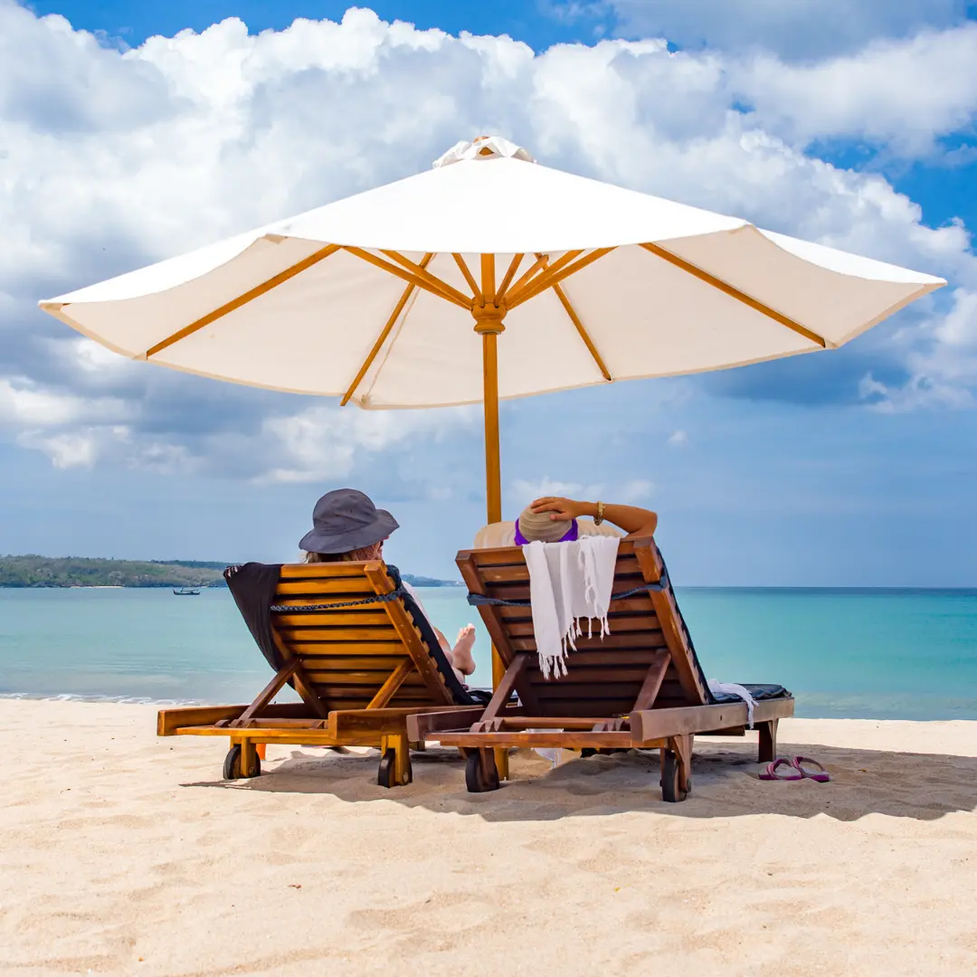 Two people relaxing on sun loungers under a large white umbrella on a sandy tropical beach facing a calm blue ocean.